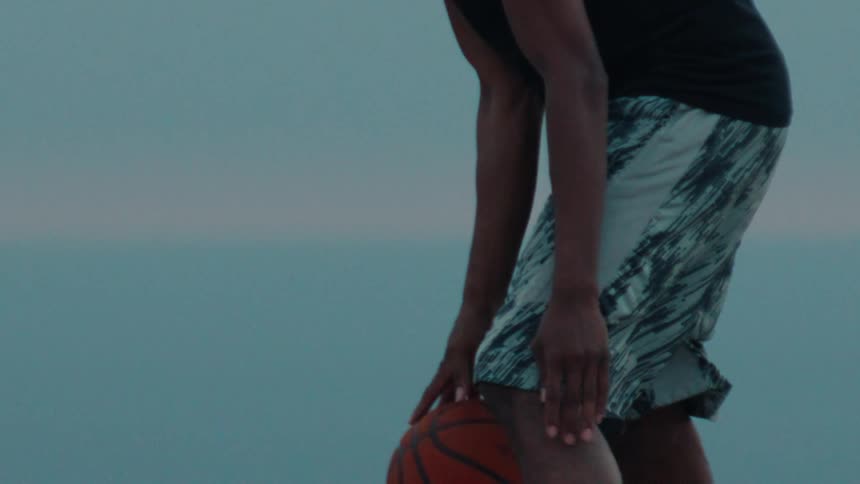 Close-up of a basketball player dribbling a ball in dramatic slow motion with an ocean background at dusk