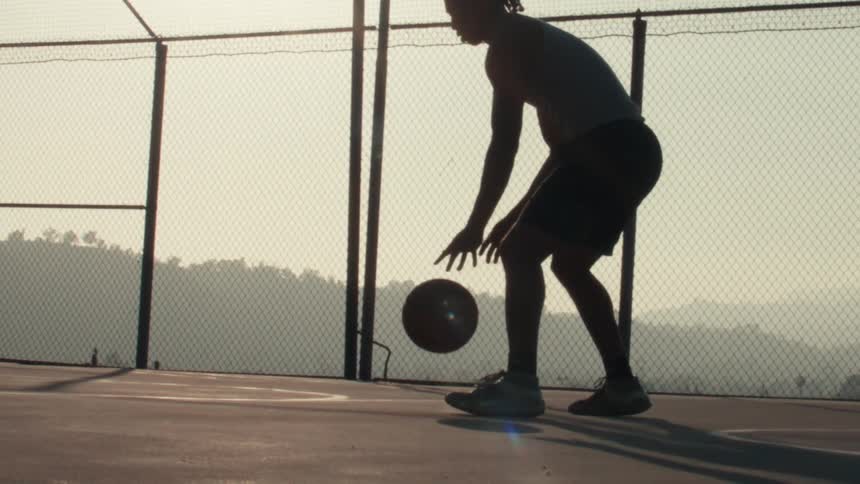 In dramatic slow motion, a silhouetted basketball player dribbles the ball on an outdoor court against the hazy morning sun
