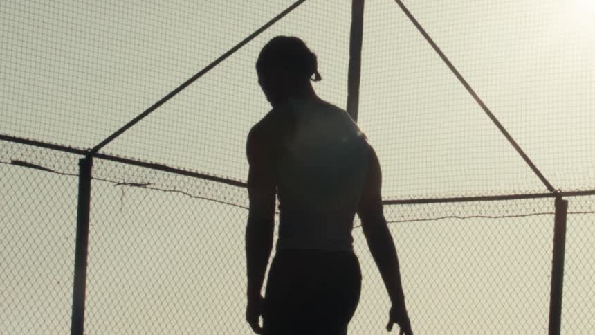 Slow motion silhouette of a basketball player dribbling and contemplating on a fenced court