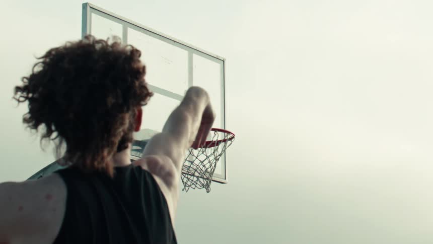 A man with curly hair shoots and scores a perfect basket in super slow motion against a bright sky