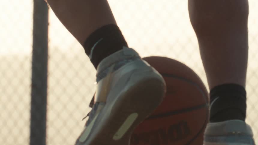 A basketball player's legs and feet dribble a ball in slow motion on an outdoor court at sunset