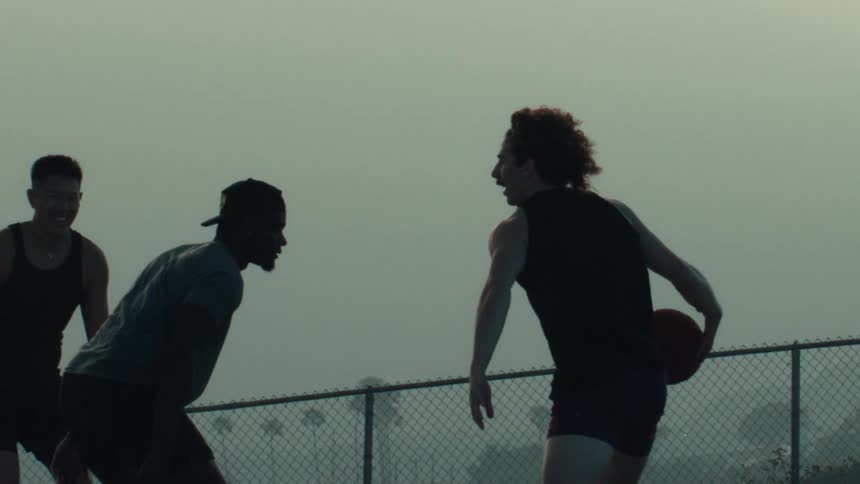 Three athletic men play a game of one-on-one basketball in slow motion, silhouetted against a hazy sky at an outdoor court