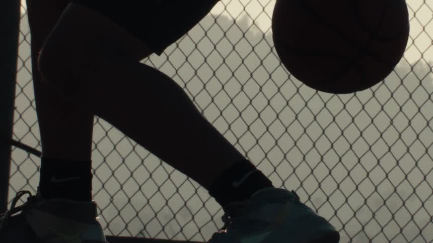 Slow motion silhouette of a basketball player's legs and hands dribbling on an outdoor court at sunrise