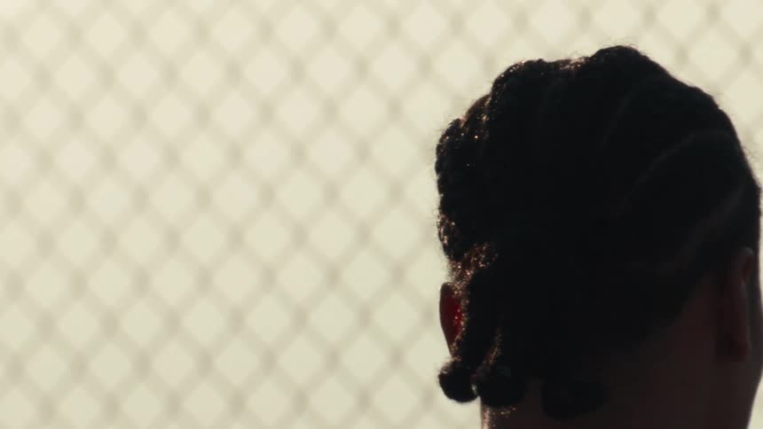Slow-motion close-up shot from behind a silhouetted basketball player with cornrows holding a ball next to a chain-link fence