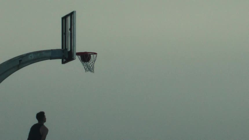 In a dramatic ultra slow-motion shot, a silhouetted basketball player scores a layup against an overcast sky