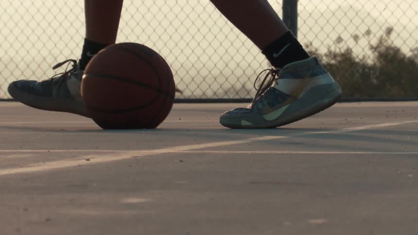 Close up of a basketball player dribbling a ball in slow motion on an outdoor court during sunset