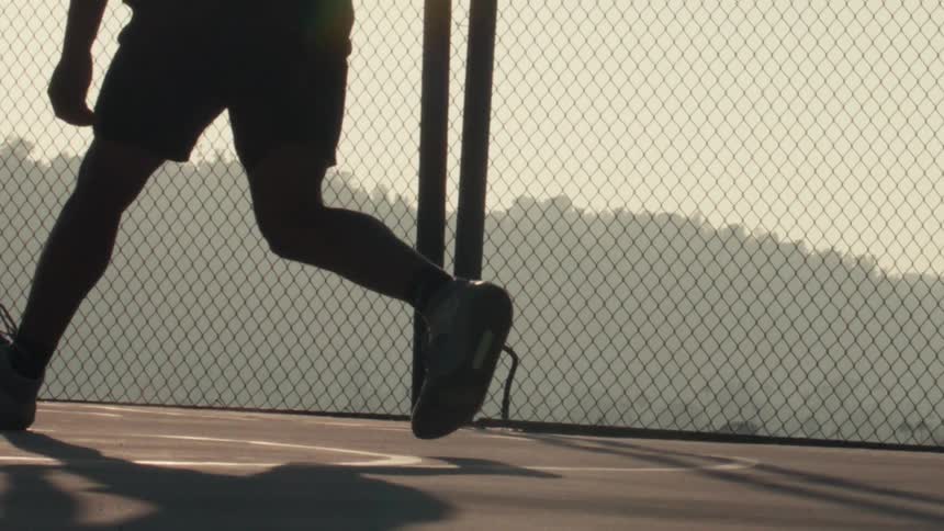 Slow motion silhouette of a basketball player dribbling on an outdoor court against a chain-link fence at sunrise