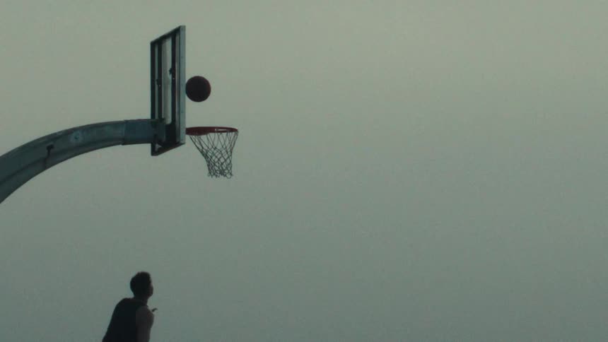 A silhouetted basketball player jumps to make a layup in super slow motion against an overcast sky