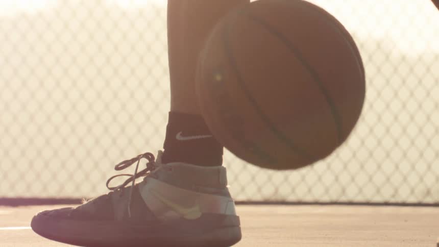 A close-up, slow-motion shot shows a basketball player's sneakers as they dribble the ball on an outdoor court during a golden sunset