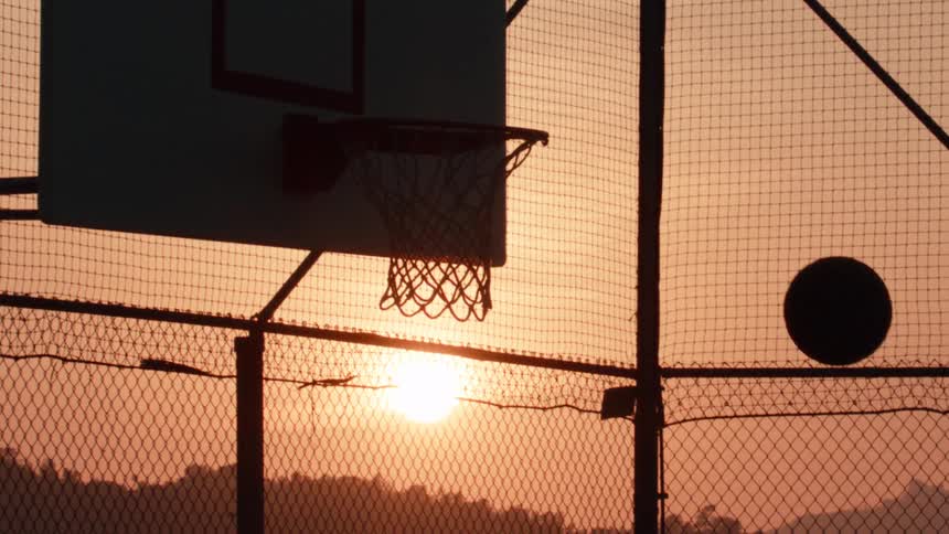 A basketball bounces off the rim in slow motion, silhouetted against the warm glow of the setting sun