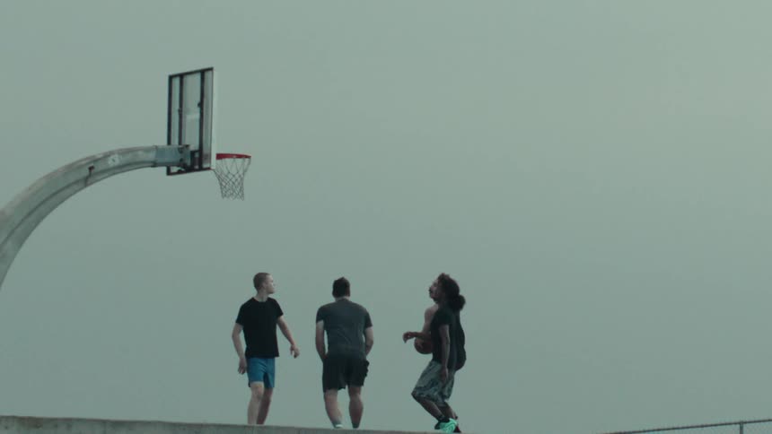 A player shoots a basketball towards the hoop in super slow motion as three other players watch on an outdoor court