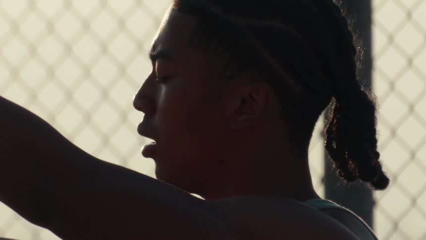 A slow-motion close-up of a silhouetted young athlete stretching against a chain-link fence during a golden hour sunset