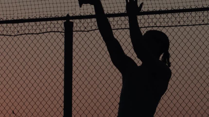 A silhouetted man slowly raises his arms in the air while standing behind a chain-link fence at sunset