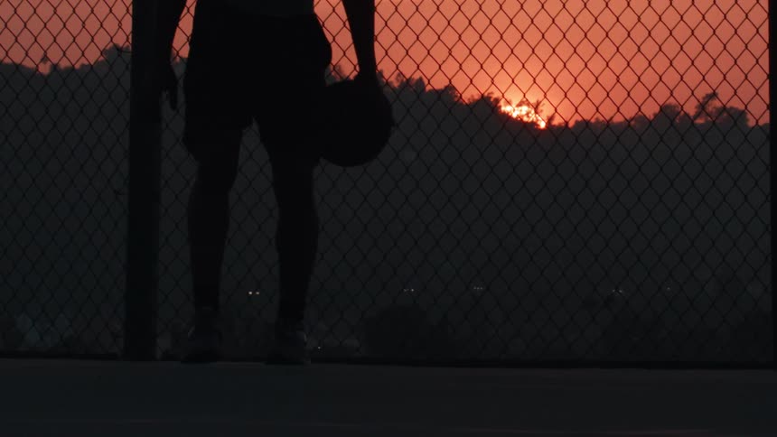 A basketball player dribbles a ball in slow motion, silhouetted against a vibrant sunset on an outdoor court