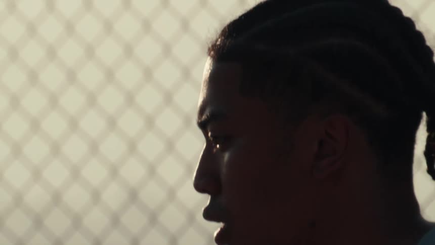 Slow-motion close-up profile of a young athlete with braided hair catching his breath against a chain-link fence at sunset