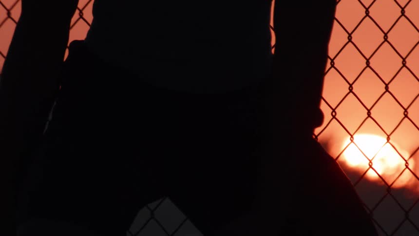 Close-up reveals the silhouette of a pensive basketball player behind a chain-link fence at sunset