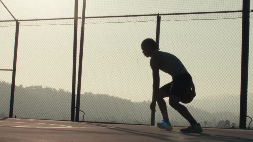 A silhouetted basketball player practices dribbling moves in super slow motion on a fenced-in court with a hazy mountain backdrop