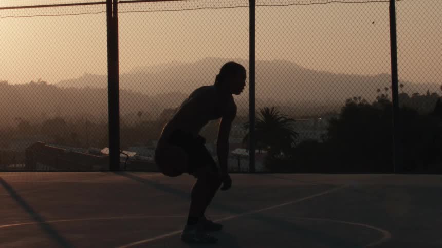Slow motion silhouette of a focused basketball player dribbling on a city court during a hazy golden sunset