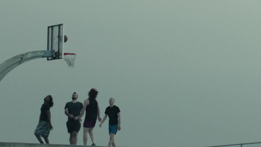 A basketball player makes an epic layup in super slow motion as his friends watch under a moody, overcast sky