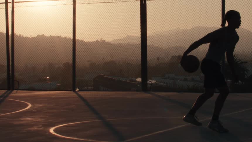 In a cinematic slow-motion silhouette, a basketball player practices dribbling on a city court as the sun sets behind distant mountains