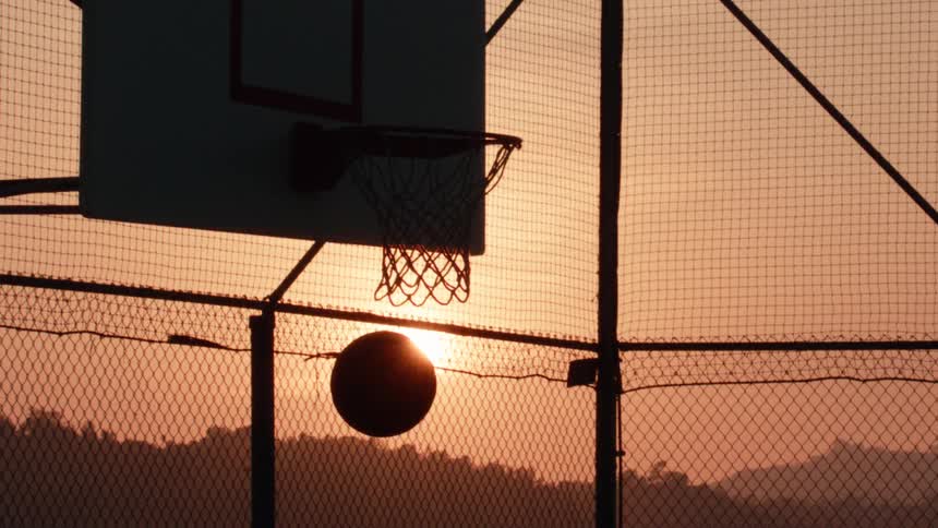 A silhouetted basketball dramatically misses the hoop in slow motion on an outdoor court as the sun sets