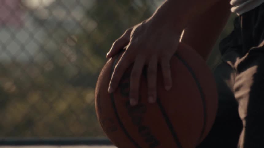 A basketball player's hands move in slow motion over a basketball on an outdoor court at sunset