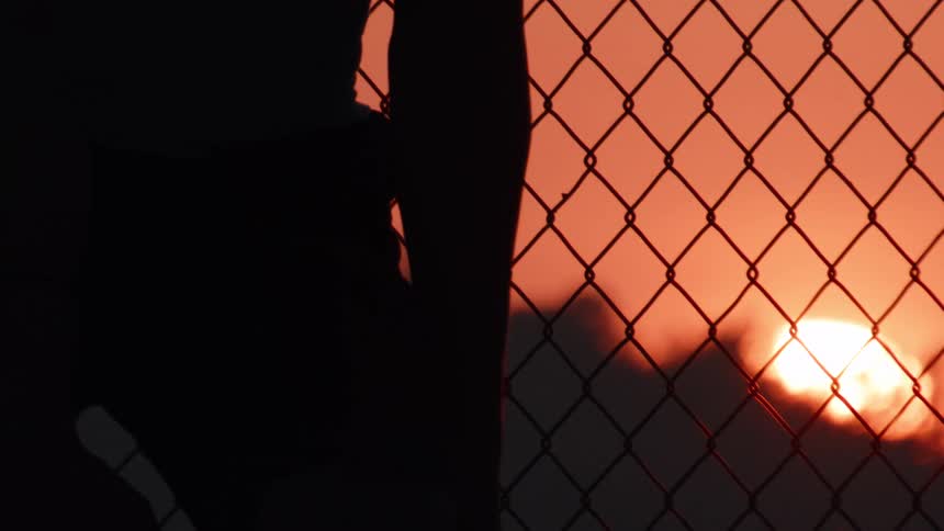 A basketball player's silhouette against a chain-link fence as they hold a basketball during a beautiful sunset