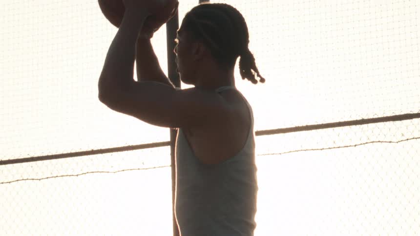 A silhouetted basketball player in slow motion aims and prepares to shoot the ball on an outdoor court with the sun flaring brightly behind him
