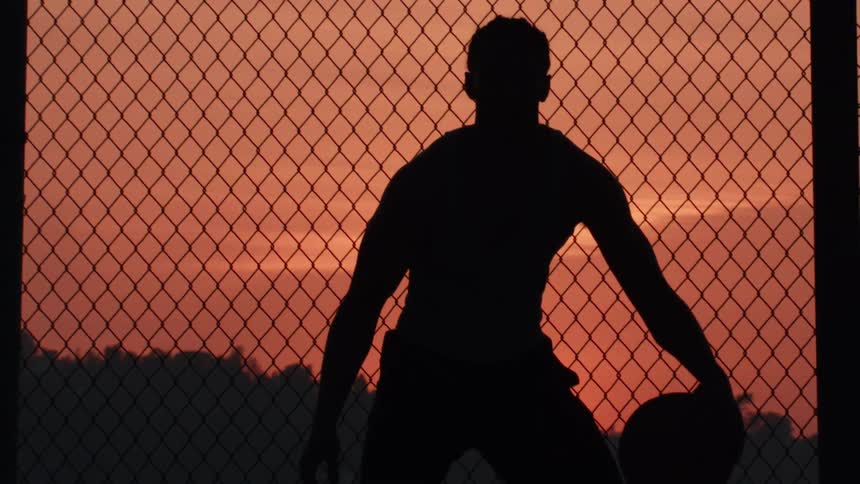 A basketball player dribbles in slow motion, silhouetted against a chain-link fence during a vibrant sunset