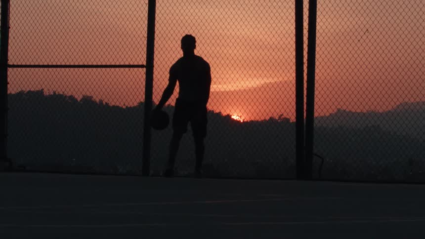 Silhouette of a man with a basketball standing on an outdoor court against a beautiful sunset over the mountains