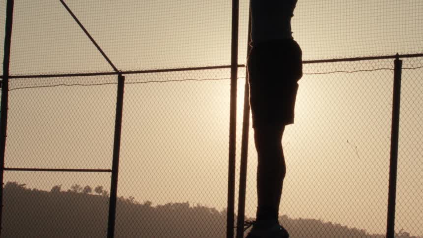 A woman performs a slow-motion squat to an overhead press with a medicine ball, silhouetted against the setting sun