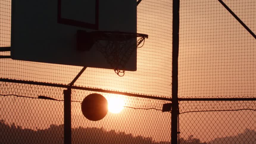 A basketball swishes through a hoop in slow motion, silhouetted against a beautiful sunset