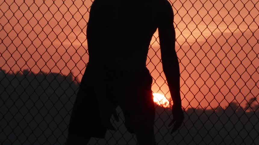 Slow motion silhouette of a basketball player holding a ball at sunset on an outdoor court behind a chain link fence