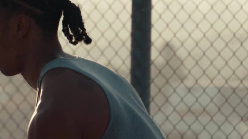 Intense slow-motion close-up of a young athlete with cornrows taking a breath against a chain-link fence at sunset