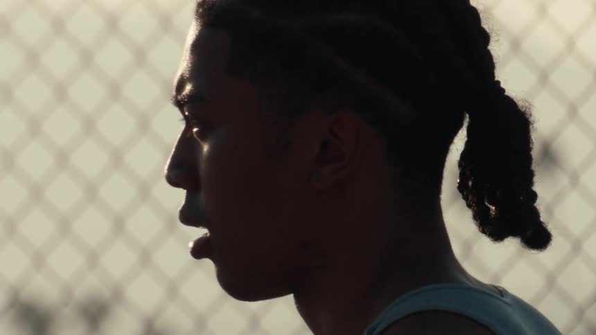 Close-up slow-motion of a determined young athlete catching his breath in profile against a chain-link fence