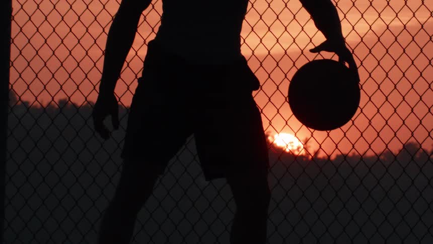 Silhouette of a person dribbling a basketball in slow motion in front of a chain-link fence during a vibrant sunset
