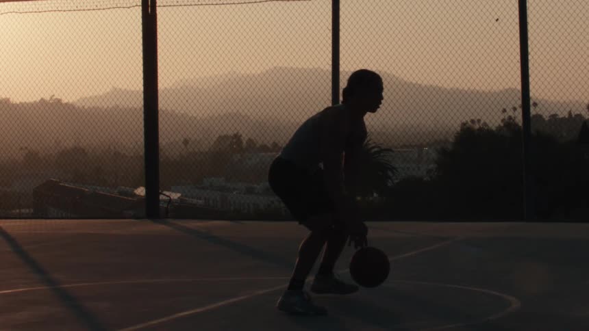 Cinematic slow motion of a silhouetted basketball player dribbling on an outdoor court with a mountain view at sunset