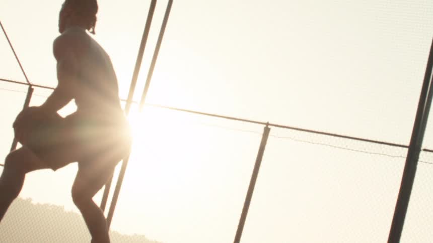 Slow zoom out reveals a silhouetted basketball player holding the ball in a ready stance on an outdoor court at sunset