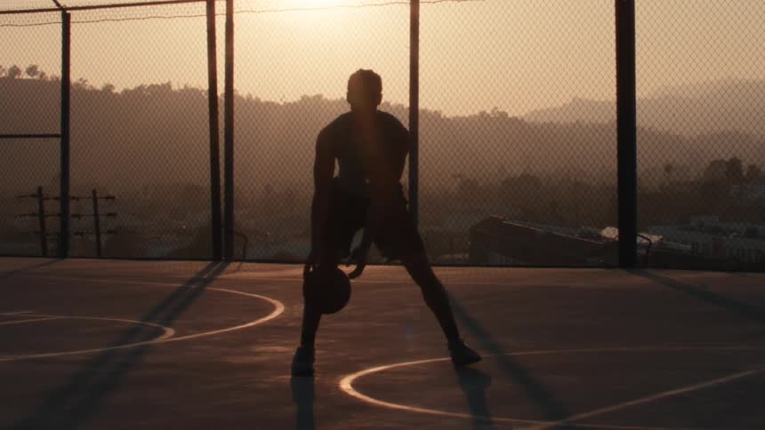 Slow motion silhouette of an athletic man dribbling a basketball on a city court during a golden sunset