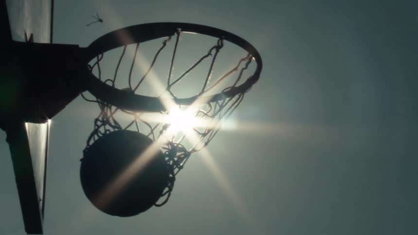 A basketball swishes through the net in slow motion, silhouetted against the bright sun creating a beautiful lens flare