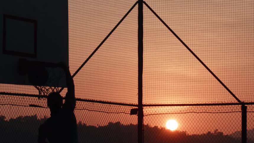 A basketball player's silhouette performs a dramatic slam dunk in slow motion against the backdrop of a beautiful sunrise