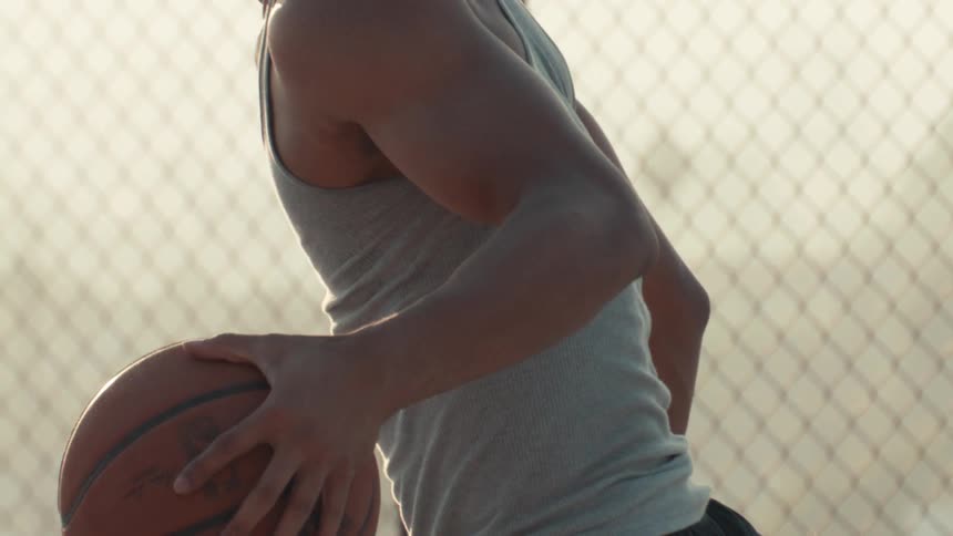 A determined young athlete with braided hair holds a basketball in slow motion on an outdoor court, backlit by the warm sunset