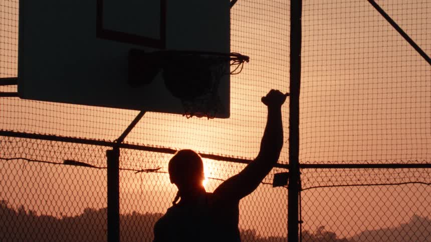 In an epic slow-motion shot, a silhouetted player gracefully slam dunks a basketball as the sun sets
