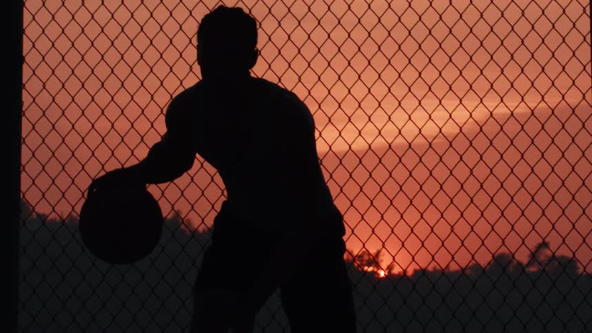 A silhouetted man dribbles a basketball in slow motion in front of a chain-link fence at sunset