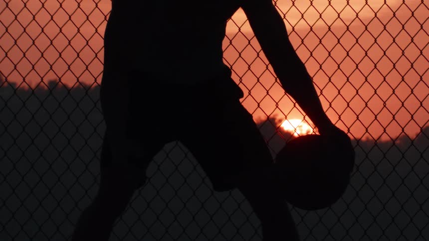 A basketball player's silhouette is seen dribbling in slow motion against a fiery sunset behind a chain-link fence