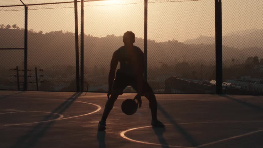 A basketball player's silhouette is captured in slow motion as they dribble on an outdoor court during a hazy golden sunset