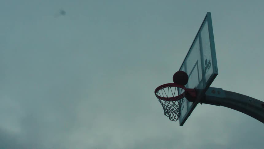 A basketball player's silhouette is captured in dramatic slow motion as they execute a powerful slam dunk against a moody, overcast sky