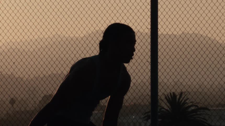 A silhouetted basketball player practices dribbling in slow motion on an outdoor court at sunrise