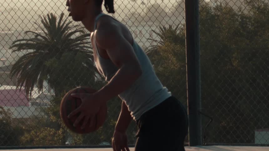 Cinematic slow motion pan reveals a young, athletic man with braided hair holding a basketball on an outdoor court at sunrise