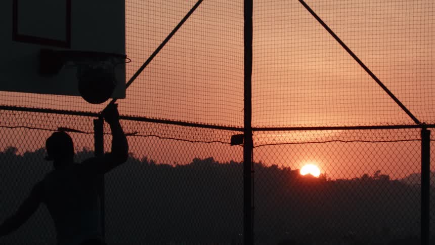 A silhouetted basketball player executes a powerful slam dunk in dramatic slow motion against a vibrant sunrise